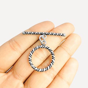 May include: Silver-toned toggle clasp with a twisted rope design on the circular ring and bar. The clasp is held in a hand, showcasing its intricate details and potential for jewelry making or craft projects.