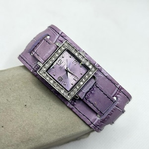 May include: A square-shaped watch with a purple leather strap and a silver face. The face is surrounded by clear rhinestones and features the word "Regal" in the centre.