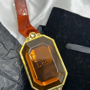 Könnte beinhalten: Ein rechteckiger Anhänger mit goldenem Rahmen und einem braunen, edelsteinartigen Mittelteil. Das Wort "Dior" ist auf dem Edelstein eingeprägt. Der Anhänger ist an einem rostfarbenen Band befestigt. Kleine, dekorative Edelsteine ​​schmücken die Ober- und Unterseite des Anhängers.