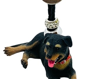 Op de afbeelding: Een hondenbedel met een zwart-bruin Rottweiler-beeldje met een rode halsband en een roze tong. De bedel hangt aan een zwart koord en een decoratieve zilveren kraal. Een ronde bronzen tag leest "My belongs to My".