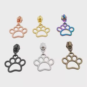 May include: Six different colored metal zipper pulls shaped like a paw print. The zipper pulls are in gold, rose gold, rainbow, black, silver, and bronze.