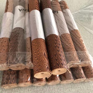 May include: A collection of rolled faux leather sheets in various shades of brown, featuring embossed textures resembling crocodile and ostrich skin. The rolls are wrapped in clear plastic, ready for use in crafting or upholstery projects.
