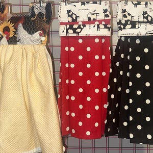 May include: Three hanging kitchen towels. The first features a rooster and hen print with a yellow textured bottom. The second is red with white polka dots, and the third is black with white polka dots. All have a loop for hanging.