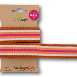 Könnte beinhalten: Braunes, weißes und rosafarbenes gestreiftes Gummiband, 200 cm lang. Das Band ist auf einer Pappekarte mit dem Text "ALB Stoffe" und "stripe 200 cm" in rosa verpackt.