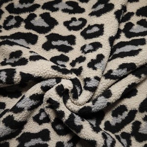 Könnte beinhalten: Nahaufnahme eines cremefarbenen Stoffes mit einem schwarzen und grauen Leopardenmuster. Das Plüschmaterial ist gefaltet und zeigt die Textur und das Design des Animal-Prints. Der Stoff eignet sich für Kleidung und Heimdekoration.