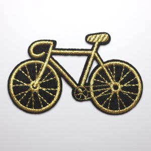 Könnte beinhalten: Gestickter Aufnäher mit einem Fahrrad-Design. Der Aufnäher hat einen schwarzen Hintergrund mit goldfarbenen Details, einschließlich Rahmen, Rädern und Lenker. Das Fahrrad ist im klassischen Rennrad-Stil gehalten.