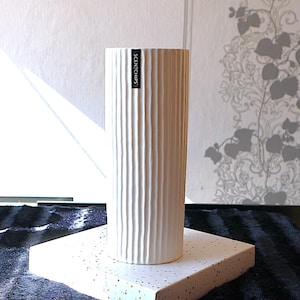 Könnte beinhalten: Eine zylindrische weiße Keramikvase mit vertikaler Rippenstruktur. Die Vase hat ein kleines schwarzes rechteckiges Etikett nahe dem oberen Rand mit dem weißen Text SCENTCHIPS. Das Objekt weist durchgehend eine glatte, matte Oberfläche auf.
