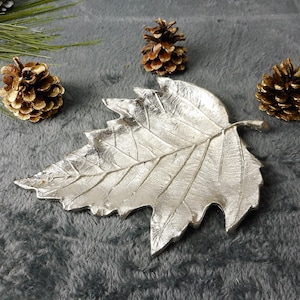 Könnte beinhalten: Eine silberne, blattförmige Servierplatte mit einem detaillierten, strukturierten Design. Die Platte ist aus Metall und hat ein natürliches, organisches Aussehen.