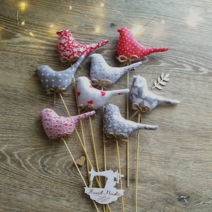 Puede incluir: Una colección de pájaros de tela hechos a mano con patrones rojos y grises sobre una superficie de madera. Los pájaros están unidos a palos de madera y tienen una etiqueta hecha a mano.