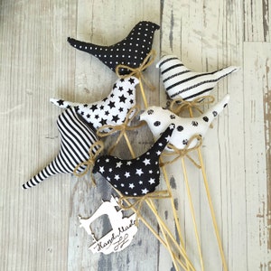 Puede incluir: Cinco pájaros de tela en blanco y negro en palos de madera. Los pájaros están decorados con lunares, rayas y estrellas. Los palos están atados con cuerda y una etiqueta que dice "Hand Made".