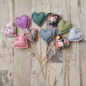 May include: Assortment of 10 heart-shaped fabric decorations with various floral and geometric patterns. The hearts are attached to wooden sticks and are arranged in a fan shape on a wooden surface.