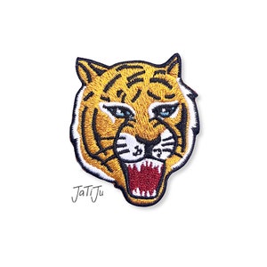 Könnte beinhalten: Gestickter Tigerkopf-Aufnäher mit grimmigem Ausdruck. Der Aufnäher hat ein gelb-schwarzes Streifendesign mit blauen Augen und einem roten, geöffneten Maul. Der Text "JaTiJu" ist in schwarzer Schreibschrift geschrieben.
