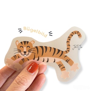 Könnte beinhalten: Ein transparenter Aufkleber mit einem Zeichentricktiger mit orangefarbenen und schwarzen Streifen, der lächelt und seine Pfoten ausstreckt. Der Aufkleber trägt den Text "Bügelbild" und den Markennamen "JatiJu".