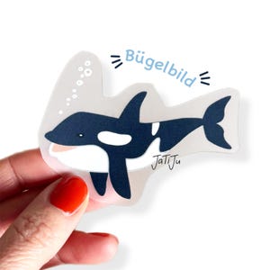 Könnte beinhalten: Ein Aufkleber mit einem Cartoon-Orca-Wal in Marineblau und Weiß, mit dem Text "Bügelbild" in Hellblau. Der Aufkleber hat einen transparenten Hintergrund und die Signatur des Künstlers "JatiJu".