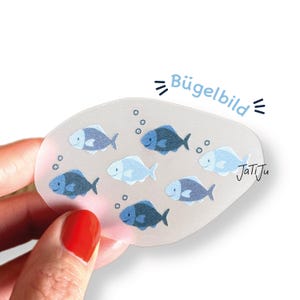 Könnte beinhalten: Ein klarer, ovaler Aufbügler mit sieben Cartoon-Fischen in Blau- und Weißtönen, mit Blasen. Der Text "Bügelbild" steht über den Fischen, und "JatiJu" ist rechts geschrieben.