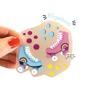Könnte beinhalten: Ein Aufkleber mit zwei Rollschuh-Illustrationen, einer hellblauen und einer rosa, jeweils mit einem Regenbogen. Der Aufkleber hat den Text "Bügelbild" und "JatiJu". Der Hintergrund ist hellbraun mit blauen, rosa und gelben Kreisen.