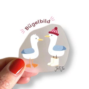 Könnte beinhalten: Ein transparenter Aufkleber mit zwei Cartoon-Möwen. Eine Möwe trägt eine rot-rosa Mütze und weiße Stiefel. Die andere Möwe hat blaue Flügel. Der Aufkleber trägt den Text "Bügelbild" und die Signatur des Künstlers.