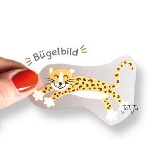 Könnte beinhalten: Ein gelb-brauner Leopard-Aufnäher mit dem Text "Bügelbild". Der Leopard lächelt und hat schwarze Flecken. Der Aufnäher befindet sich auf einem grauen Hintergrund.