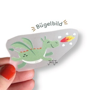 Könnte beinhalten: Ein grüner Cartoon-Drachen-Bügelbild mit einem lächelnden Gesicht, Flügeln und Feueratem. Das Bügelbild befindet sich auf einem transparenten Hintergrund mit dem Text "Bügelbild" und der Signatur des Künstlers "JatiJu".