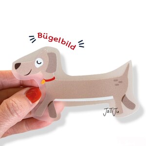 Könnte beinhalten: Hellbrauner Cartoon-Hund-Aufkleber mit rotem Halsband und kleinem gelben Anhänger. Der Aufkleber hat den Text "Bügelbild" in Rot und die Signatur des Künstlers "JatiJu".