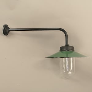 Op de afbeelding: Zwarte wandlamp met een groene metalen kap en een heldere glazen bol. De lamp heeft een lange, gebogen arm.
