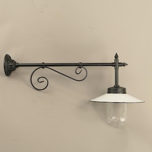 Può includere: Applique da parete per esterni in metallo nero. L'apparecchio ha un paralume bianco rivolto verso il basso con finiture nere e un involucro di vetro trasparente. Il braccio decorativo presenta un design a volute.