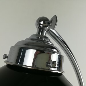May include: Close-up of a chrome desk lamp with a black shade. The lamp has a round base and a curved arm. The lamp is adjustable and has a knob on the side to control the light.