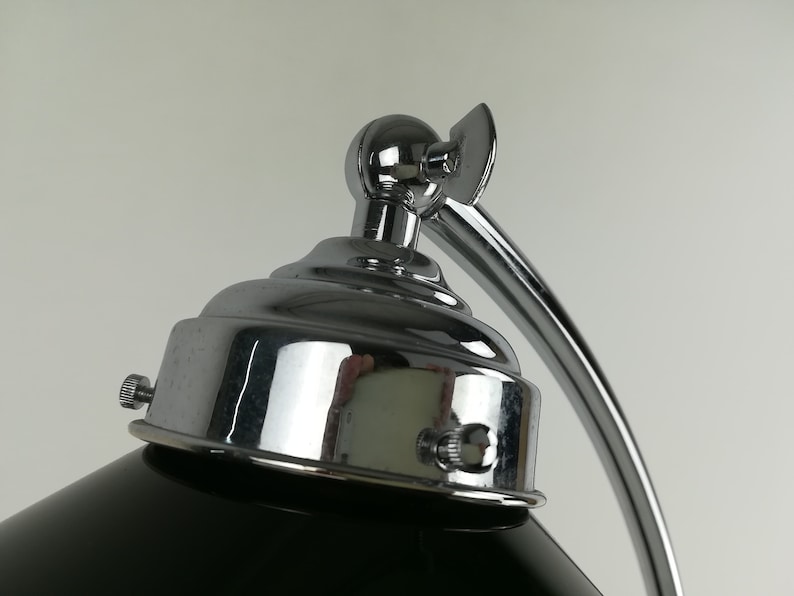May include: Close-up of a chrome desk lamp with a black shade. The lamp has a round base and a curved arm. The lamp is adjustable and has a knob on the side to control the light.