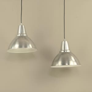 May include: Two silver-colored pendant lights with a dome-shaped shade and a white interior. The lights are suspended from black cords against a neutral-toned wall. The design is industrial and minimalist.