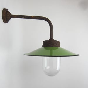 Pode incluir: Uma luminária de parede antiga com um braço castanho enferrujado e uma sombra de esmalte verde. A sombra tem uma borda preta e um globo de vidro transparente. A luminária é montada em uma parede branca.