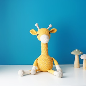 May include: A handmade yellow crochet giraffe with white accents, sitting on a white surface against a blue background. Two wooden mushroom decorations are next to the giraffe. The giraffe has white spots and a white muzzle.