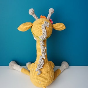 May include: A handmade, yellow crocheted giraffe toy with gray and white accents. The giraffe has a long neck, small ears, and a tail. It is decorated with small flowers and sits on a white surface against a blue background.