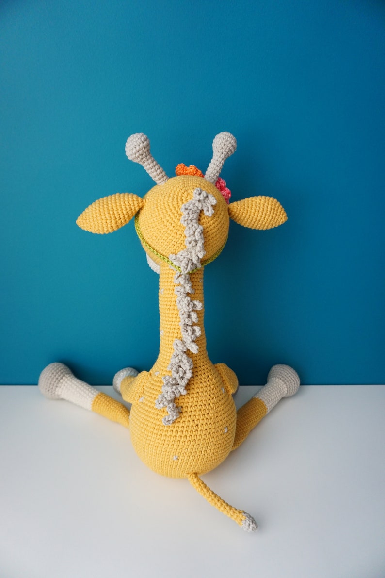 May include: A handmade, yellow crocheted giraffe toy with gray and white accents. The giraffe has a long neck, small ears, and a tail. It is decorated with small flowers and sits on a white surface against a blue background.