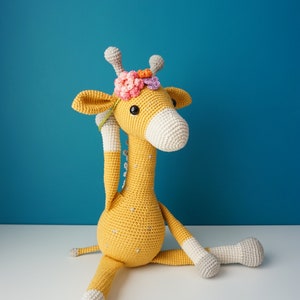 May include: A handmade crochet giraffe in yellow, white, and gray, adorned with a floral crown. The plush toy has a long neck, large eyes, and is sitting with its legs extended. The giraffe measures approximately 20 inches tall.