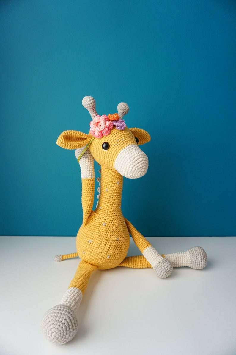 May include: A handmade crochet giraffe in yellow, white, and gray, adorned with a floral crown. The plush toy has a long neck, large eyes, and is sitting with its legs extended. The giraffe measures approximately 20 inches tall.