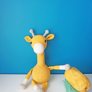 May include: A handmade, yellow crochet giraffe with white accents and small white dots. The giraffe is sitting on a white surface against a blue background. A ball of yellow yarn and a ball of green yarn are next to the giraffe.