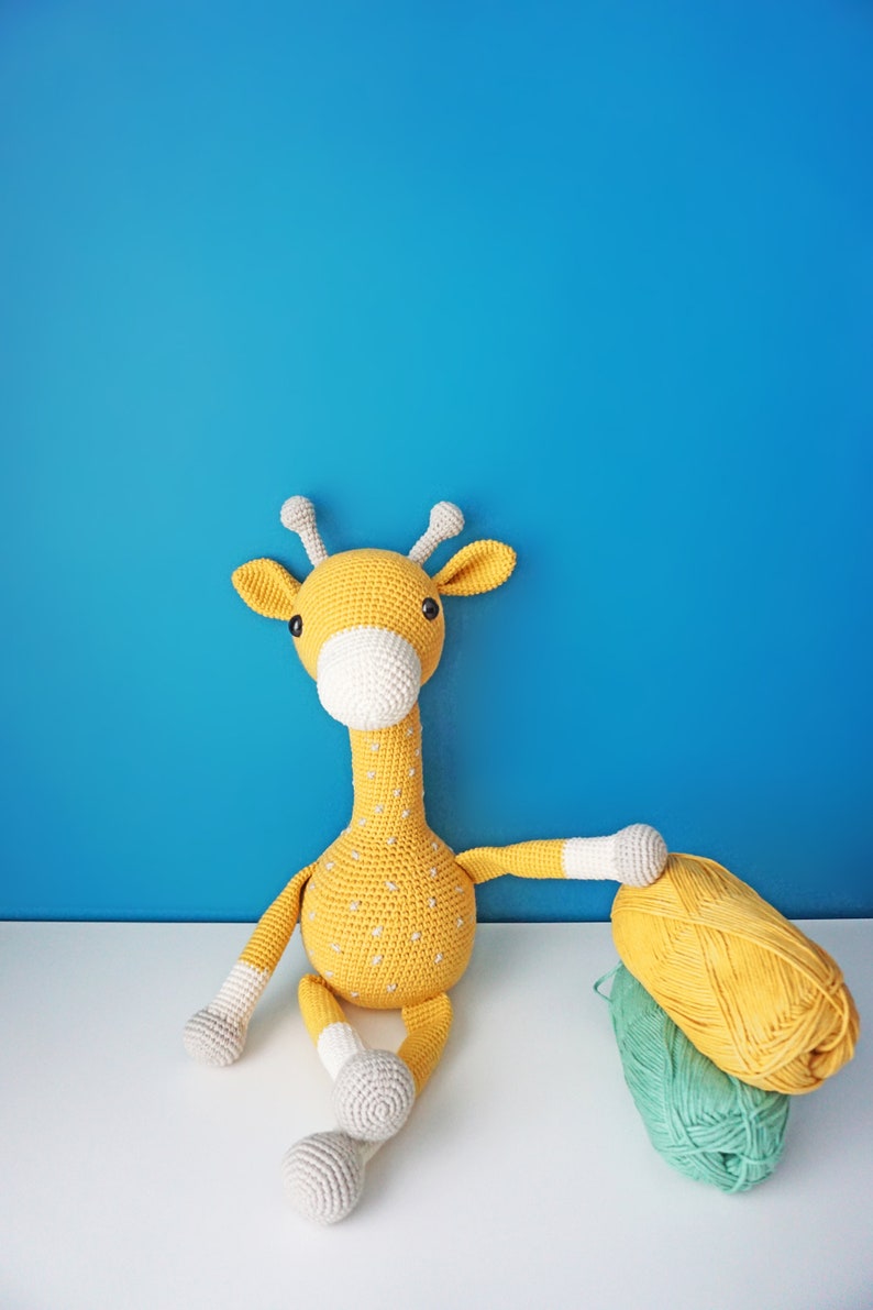May include: A handmade, yellow crochet giraffe with white accents and small white dots. The giraffe is sitting on a white surface against a blue background. A ball of yellow yarn and a ball of green yarn are next to the giraffe.