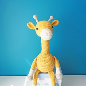 May include: A handmade, crocheted giraffe toy in yellow with white accents. The giraffe has a long neck, small horns, and is sitting on a box with a blue and white cloud pattern. The background is a solid blue.