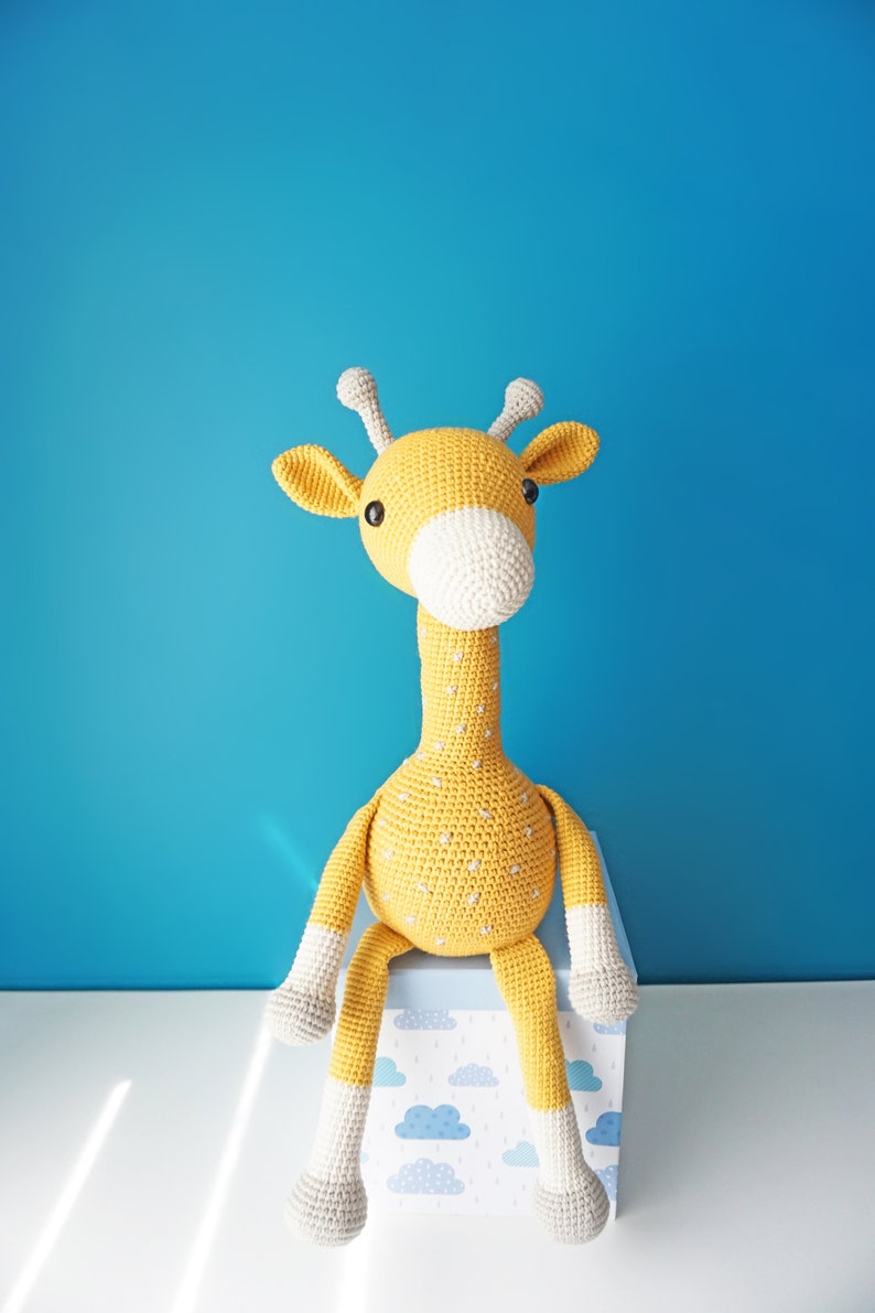 May include: A handmade, crocheted giraffe toy in yellow with white accents. The giraffe has a long neck, small horns, and is sitting on a box with a blue and white cloud pattern. The background is a solid blue.