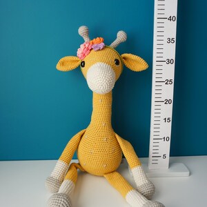 May include: A handmade, crochet giraffe toy in yellow, white, and gray, with a floral headband. The giraffe is sitting next to a white height chart. The giraffe's height is approximately 30 inches.