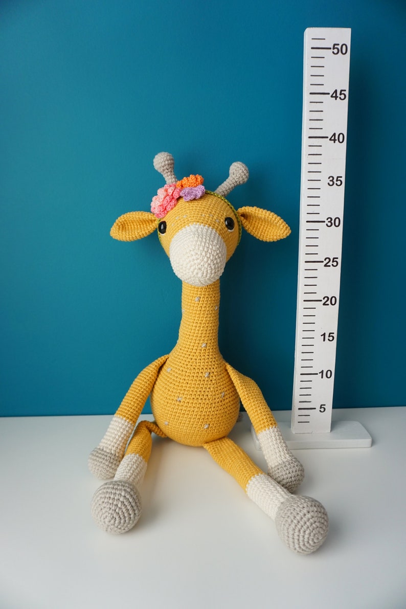 May include: A handmade, crochet giraffe toy in yellow, white, and gray, with a floral headband. The giraffe is sitting next to a white height chart. The giraffe's height is approximately 30 inches.