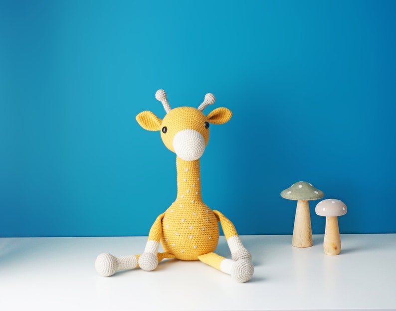 May include: A handmade yellow crochet giraffe with white accents, sitting on a white surface against a blue background. Two wooden mushroom decorations are next to the giraffe. The giraffe has white spots and a white muzzle.