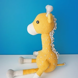 May include: A handmade crochet giraffe toy in yellow, white, and gray. The giraffe has a long neck, small horns, and is sitting down. The toy has small gray star-shaped details on its body and is set against a blue background.