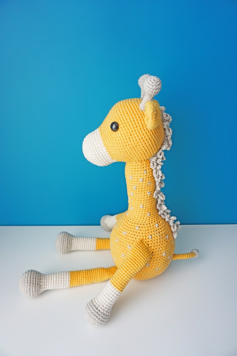 May include: A handmade crochet giraffe toy in yellow, white, and gray. The giraffe has a long neck, small horns, and is sitting down. The toy has small gray star-shaped details on its body and is set against a blue background.
