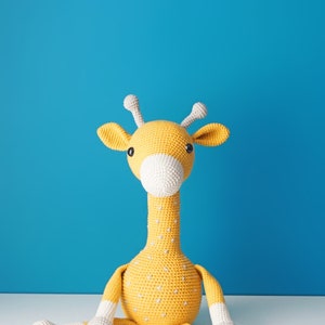 May include: A handmade, crocheted giraffe toy in yellow with white accents. The giraffe has a long neck, small horns, and a white muzzle. It is sitting on a white surface against a blue background. The toy is approximately 12 inches tall.