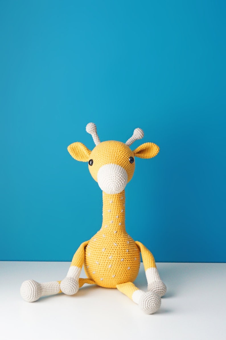 May include: A handmade, crocheted giraffe toy in yellow with white accents. The giraffe has a long neck, small horns, and a white muzzle. It is sitting on a white surface against a blue background. The toy is approximately 12 inches tall.