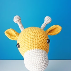 May include: A handmade crochet giraffe toy with a yellow body, white muzzle, and gray horns. The giraffe has black safety eyes and is set against a blue background. A cute and cuddly stuffed animal.