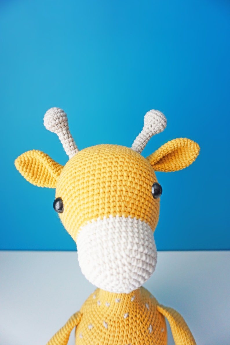 May include: A handmade crochet giraffe toy with a yellow body, white muzzle, and gray horns. The giraffe has black safety eyes and is set against a blue background. A cute and cuddly stuffed animal.