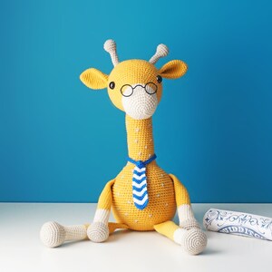 May include: A handmade yellow crochet giraffe wearing glasses and a blue and white striped tie. The giraffe has white accents on its ears, muzzle, and legs. A rolled-up scroll with blue and white designs is next to the giraffe.