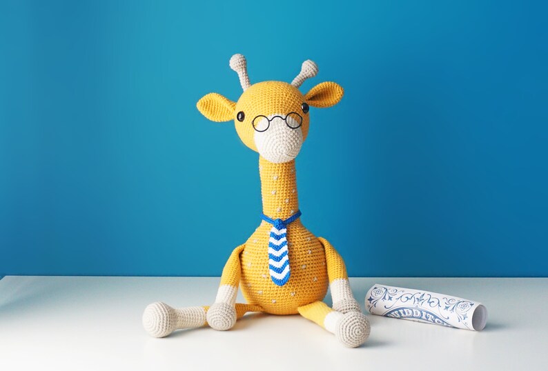 May include: A handmade yellow crochet giraffe wearing glasses and a blue and white striped tie. The giraffe has white accents on its ears, muzzle, and legs. A rolled-up scroll with blue and white designs is next to the giraffe.
